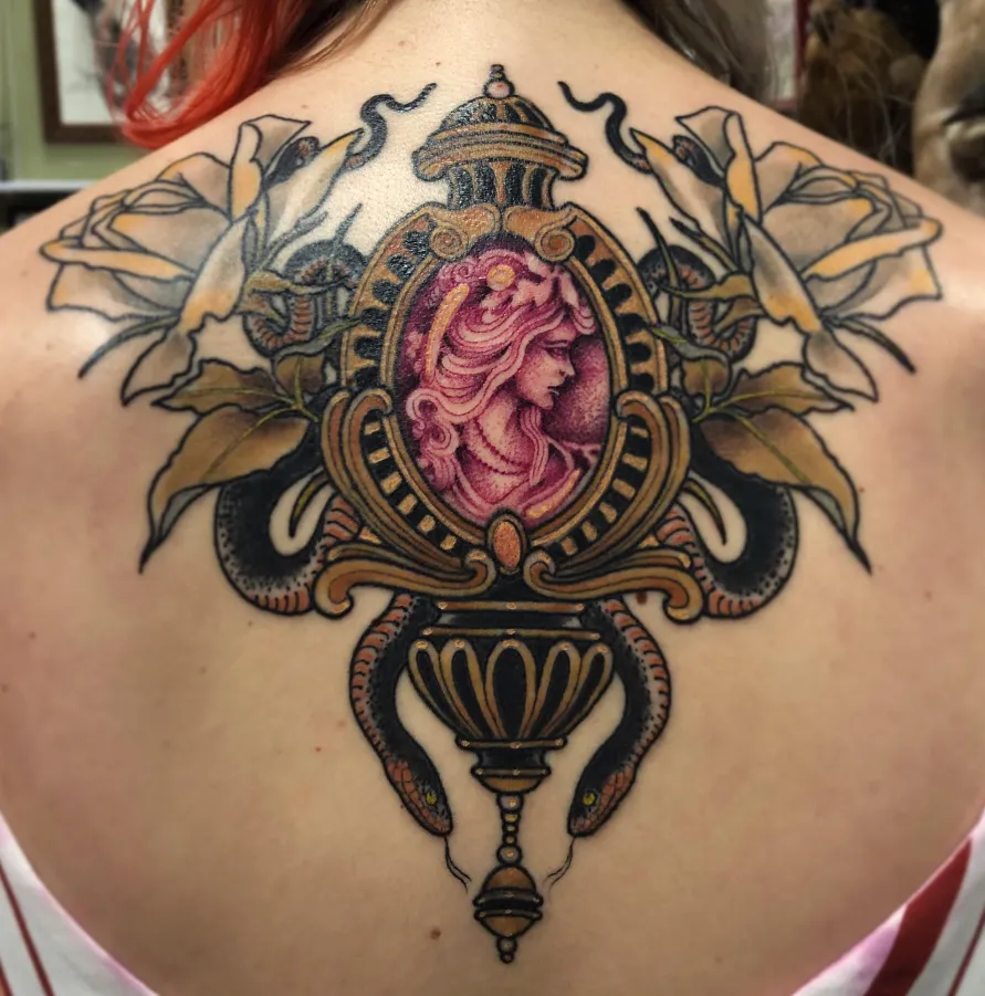 Ornate heraldic back tattoo with a pink cameo centrepiece framed in gold scrollwork and pale botanicals.