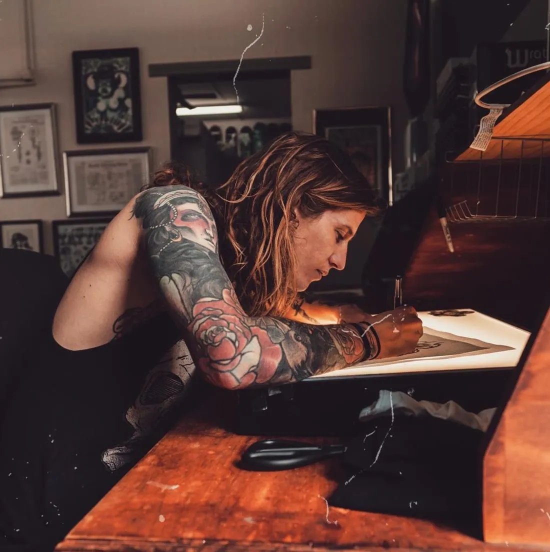 Tom Bartley leaning over a lightbox drawing a tattoo design, tattooed arms with a neo-traditional rose sleeve, in a studio hung with framed art.